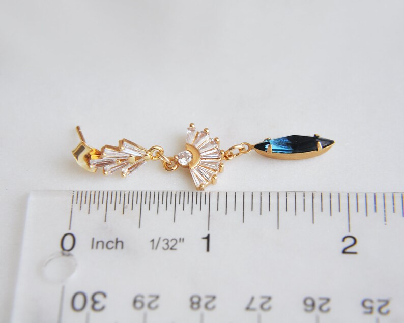 May include: A gold-toned earring with a fan-shaped design of clear crystals and a dangling blue and black crystal. The earring is approximately 2 inches long, as measured by the ruler in the image. The earring has a post back.