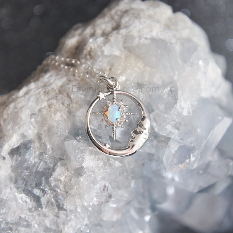 Puede incluir: Un collar de plata con un dise&ntilde;o celestial. El colgante presenta una luna creciente, una estrella y una estrella central con un &oacute;palo de color claro. El collar se muestra sobre un fondo azul claro texturizado.