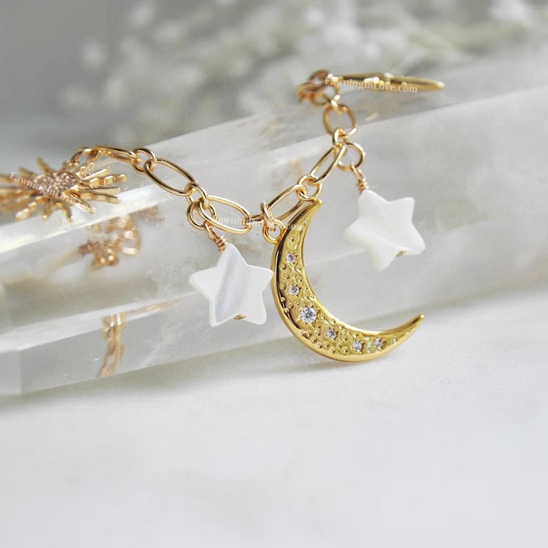 May include: Gold-toned celestial charm bracelet featuring a crescent moon pendant with embedded crystals, two white star charms, and a sunburst charm. The bracelet is linked with a delicate chain, creating a whimsical and elegant accessory.