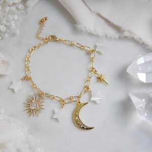 May include: Gold-toned charm bracelet featuring celestial-themed charms. The bracelet includes a sunburst charm with a central crystal, a crescent moon, star charms, and white star-shaped charms. The bracelet is displayed on a white surface.