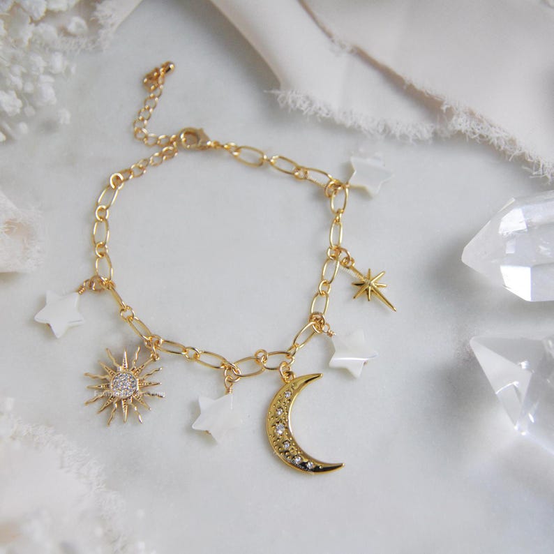 May include: Gold-toned charm bracelet featuring celestial-themed charms. The bracelet includes a sunburst charm with a central crystal, a crescent moon, star charms, and white star-shaped charms. The bracelet is displayed on a white surface.