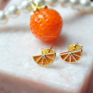 May include: Gold-tone stud earrings shaped like orange slices, each with a small, clear gem in the center. The earrings have an orange enamel filling and are set against a blurred background with an orange sphere and a string of pearls.