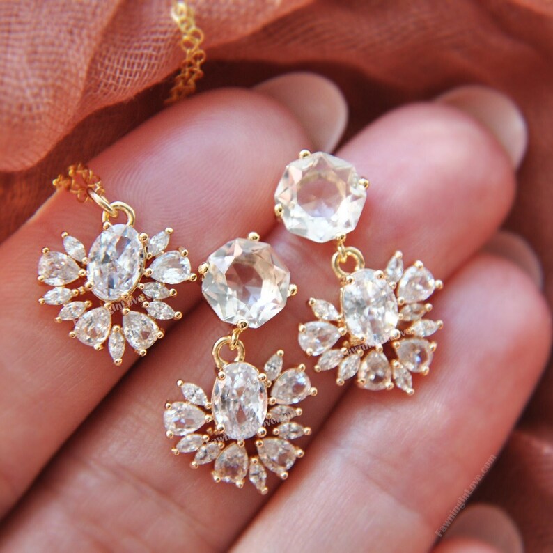 May include: A gold-toned jewelry set featuring a necklace and earrings. The necklace has a pendant with an oval-shaped clear stone surrounded by smaller clear stones. The earrings have a similar design, with a large clear stone and smaller clear stones.
