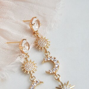 May include: Gold-toned celestial earrings featuring clear, faceted crystals and sun and crescent moon charms. The earrings have a post-back closure. The sun charms are encrusted with small, sparkling crystals. The earrings are a delicate and elegant accessory.