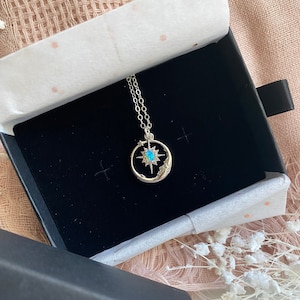 Puede incluir: Un collar plateado con un dise&ntilde;o celestial, presentado en una caja de joyer&iacute;a negra. El colgante presenta una estrella turquesa dentro de un marco circular, con estrellas y una luna creciente. La cadena del collar es delicada y plateada.