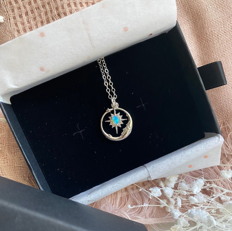Puede incluir: Un collar plateado con un dise&ntilde;o celestial, presentado en una caja de joyer&iacute;a negra. El colgante presenta una estrella turquesa dentro de un marco circular, con estrellas y una luna creciente. La cadena del collar es delicada y plateada.