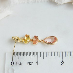 May include: A gold-toned earring with a stud post, featuring a series of colorful gemstones. The design includes a cluster of small clear stones, followed by yellow, peach, and a teardrop-shaped pink stone. The earring is approximately 2 inches long.