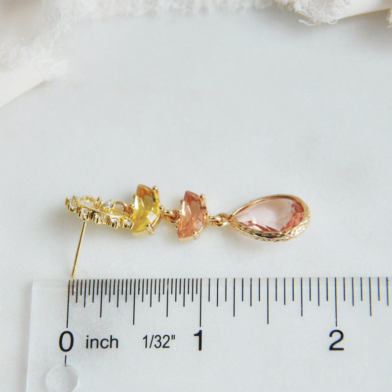 May include: A gold-toned earring with a stud post, featuring a series of colorful gemstones. The design includes a cluster of small clear stones, followed by yellow, peach, and a teardrop-shaped pink stone. The earring is approximately 2 inches long.