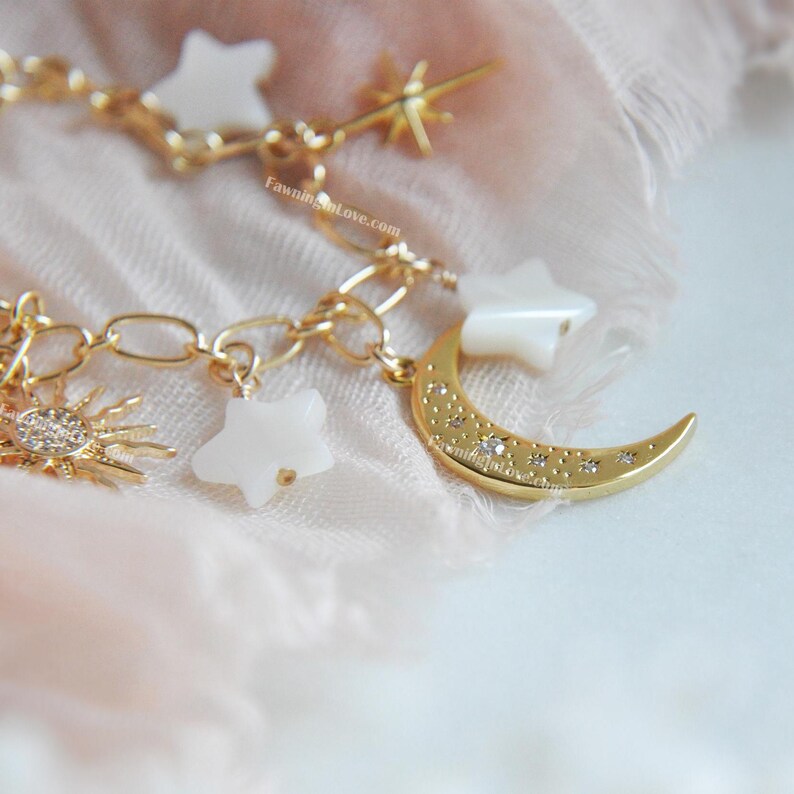 May include: A gold-colored charm bracelet featuring celestial-themed charms. The bracelet includes a crescent moon, stars, and a sun, all adorned with small crystals. The charms are suspended from a delicate chain.