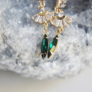 May include: Gold-toned earrings featuring a fan-shaped design with clear crystals and emerald green teardrop-shaped gems. The earrings have a delicate, elegant appearance, with a focus on the contrast between the gold metal and the vibrant green stones.