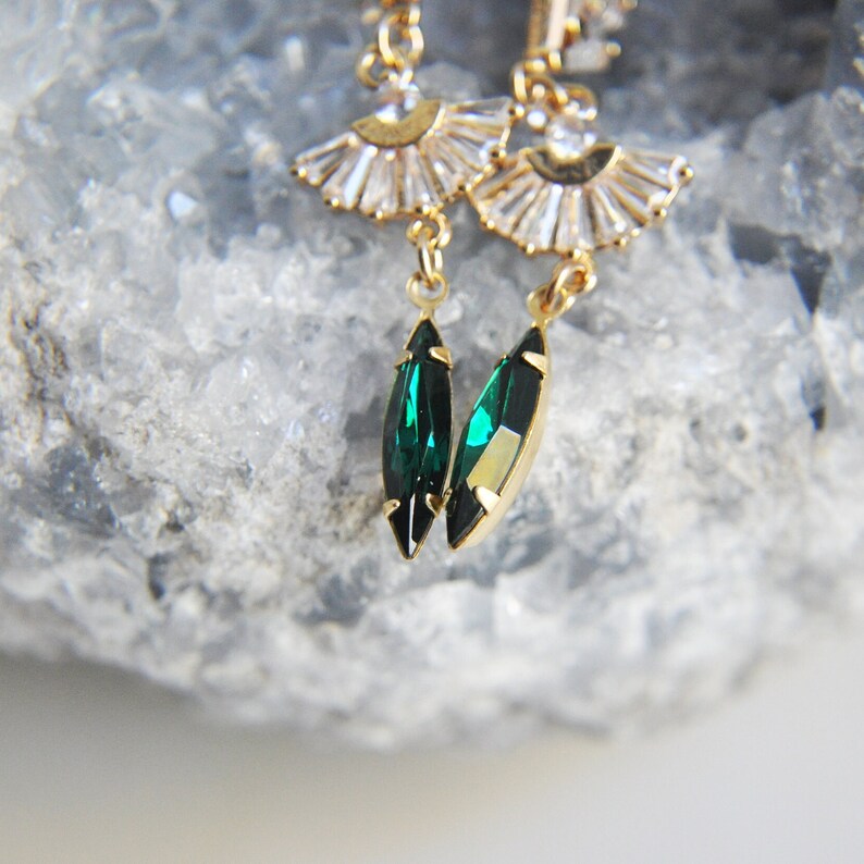May include: Gold-toned earrings featuring a fan-shaped design with clear crystals and emerald green teardrop-shaped gems. The earrings have a delicate, elegant appearance, with a focus on the contrast between the gold metal and the vibrant green stones.
