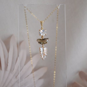 Puede incluir: Un collar dorado con tres niveles de piedras preciosas. La piedra superior es un rectángulo transparente, el centro es un semicírculo de cuarzo ahumado y la parte inferior es un grupo en forma de hoja de cristales transparentes. El collar se exhibe en un soporte transparente.