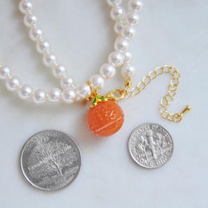 May include: A double-strand pearl necklace with a gold-toned clasp and an orange charm. The charm is a textured orange sphere with green leaves and a gold-toned bail. The necklace includes a gold-toned extender chain.
