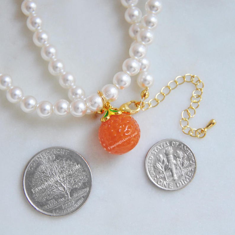 May include: A double-strand pearl necklace with a gold-toned clasp and an orange charm. The charm is a textured orange sphere with green leaves and a gold-toned bail. The necklace includes a gold-toned extender chain.