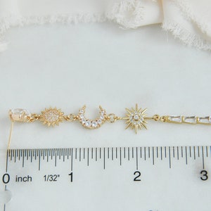 May include: Gold-toned celestial-themed earring featuring a sun, crescent moon, and starburst design. The earring has a clear, oval-shaped stud and a linear section with rectangular crystals. The earring measures approximately 3 inches in length.