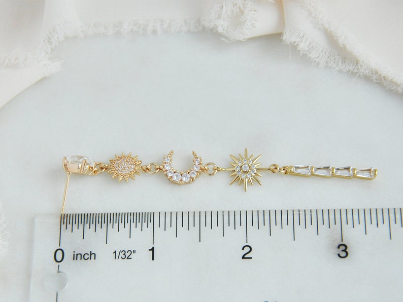 May include: Gold-toned celestial-themed earring featuring a sun, crescent moon, and starburst design. The earring has a clear, oval-shaped stud and a linear section with rectangular crystals. The earring measures approximately 3 inches in length.