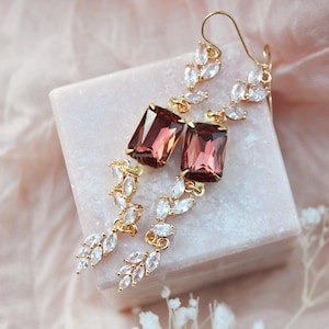 May include: Gold-toned drop earrings featuring large, rectangular, reddish-brown gemstones. The earrings have a delicate design with smaller, clear, oval-shaped stones and are displayed on a light pink surface.