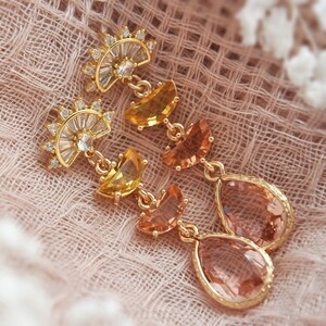 May include: Gold-toned drop earrings with a sunburst design at the top, featuring clear and yellow-toned crystals. The earrings have a teardrop-shaped, peach-colored stone at the bottom.