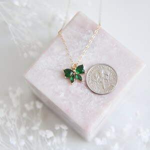 Puede incluir: Un collar dorado con un colgante en forma de lazo adornado con piedras preciosas de color verde esmeralda. El collar se exhibe sobre un bloque rectangular rosa, con una moneda para comparar el tama&ntilde;o. El fondo es blanco.