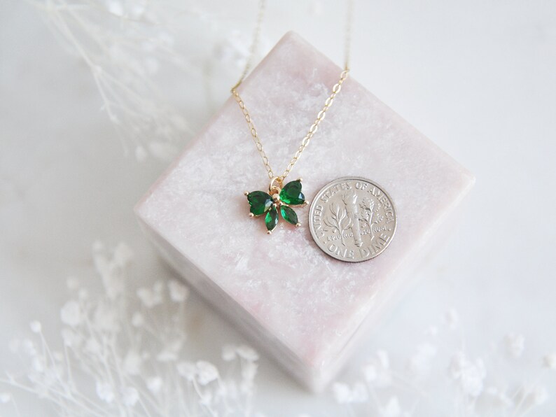 Puede incluir: Un collar dorado con un colgante en forma de lazo adornado con piedras preciosas de color verde esmeralda. El collar se exhibe sobre un bloque rectangular rosa, con una moneda para comparar el tama&ntilde;o. El fondo es blanco.