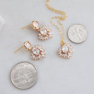 May include: A gold-toned jewelry set featuring a necklace and matching earrings. The necklace has a pendant with a central oval-shaped stone surrounded by smaller stones. The earrings have a similar design. The set is displayed on a light-colored surface.