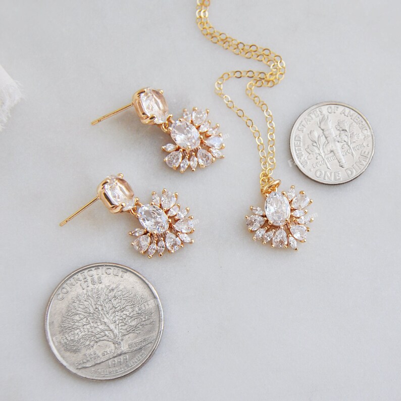 May include: A gold-toned jewelry set featuring a necklace and matching earrings. The necklace has a pendant with a central oval-shaped stone surrounded by smaller stones. The earrings have a similar design. The set is displayed on a light-colored surface.