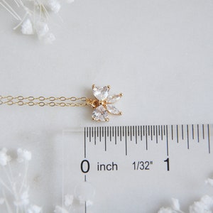 K&ouml;nnte beinhalten: Eine zarte, goldfarbene Halskette mit einem floralen Anh&auml;nger mit klaren, funkelnden Edelsteinen. Der Anh&auml;nger ist etwa 1,3 cm gro&szlig;. Die Halskette wird auf einer wei&szlig;en Oberfl&auml;che mit einem Lineal und dekorativen wei&szlig;en Blumen pr&auml;sentiert.