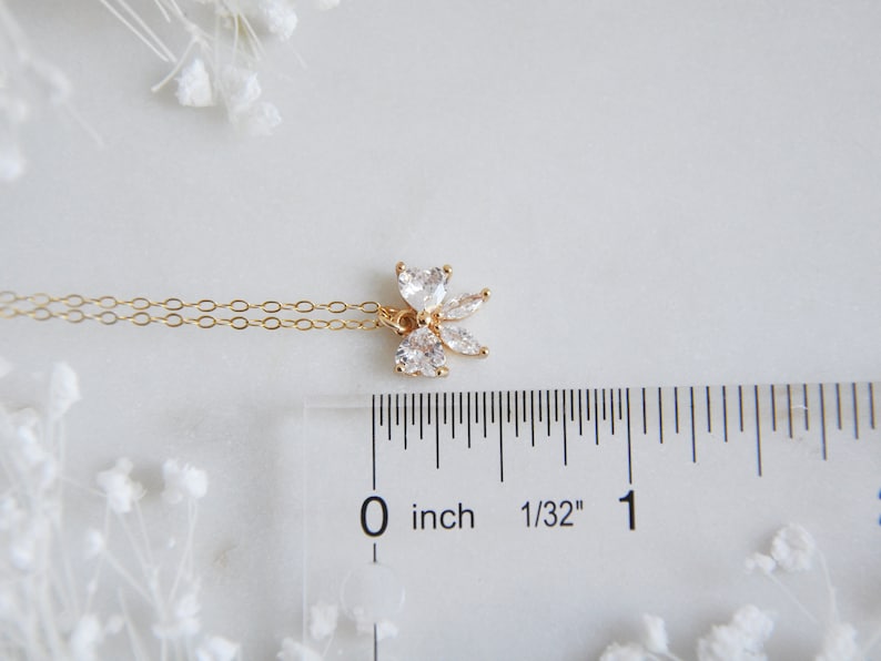 K&ouml;nnte beinhalten: Eine zarte, goldfarbene Halskette mit einem floralen Anh&auml;nger mit klaren, funkelnden Edelsteinen. Der Anh&auml;nger ist etwa 1,3 cm gro&szlig;. Die Halskette wird auf einer wei&szlig;en Oberfl&auml;che mit einem Lineal und dekorativen wei&szlig;en Blumen pr&auml;sentiert.