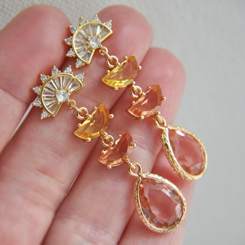 May include: Gold-toned earrings with a fan-shaped top adorned with clear crystals and a central stone. The earrings feature a series of orange and yellow-toned gemstones, culminating in a teardrop-shaped stone. The earrings are held in a hand.