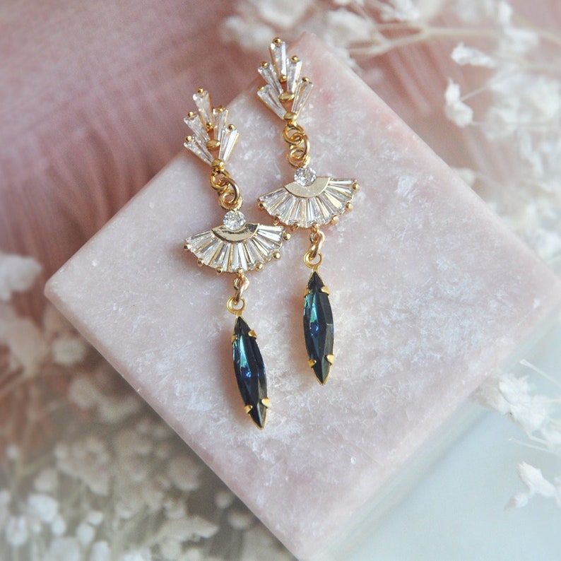 May include: Elegant gold-toned earrings with a fan-shaped design, adorned with clear crystals and a dangling dark blue teardrop-shaped stone. The earrings are displayed on a pink surface, showcasing their intricate details and sparkling appearance.