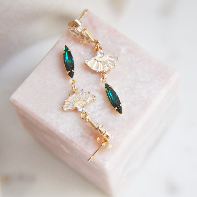 May include: Gold-toned dangle earrings featuring fan-shaped and teardrop-shaped emerald green and clear crystals. The earrings are displayed on a light pink, square-shaped surface. The earrings have a post-back closure.