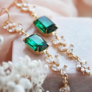 May include: Gold-toned drop earrings featuring emerald-cut green gemstones and clear, marquise-cut crystals. The earrings have a delicate, leaf-like design with a post-back closure. The jewelry is displayed on a pink and white surface.
