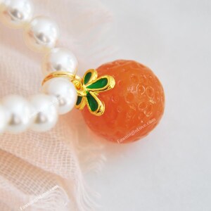 May include: A close-up of a pearl bracelet with a gold charm. The charm is an orange sphere with a gold and green accent. The bracelet is on a pink fabric.