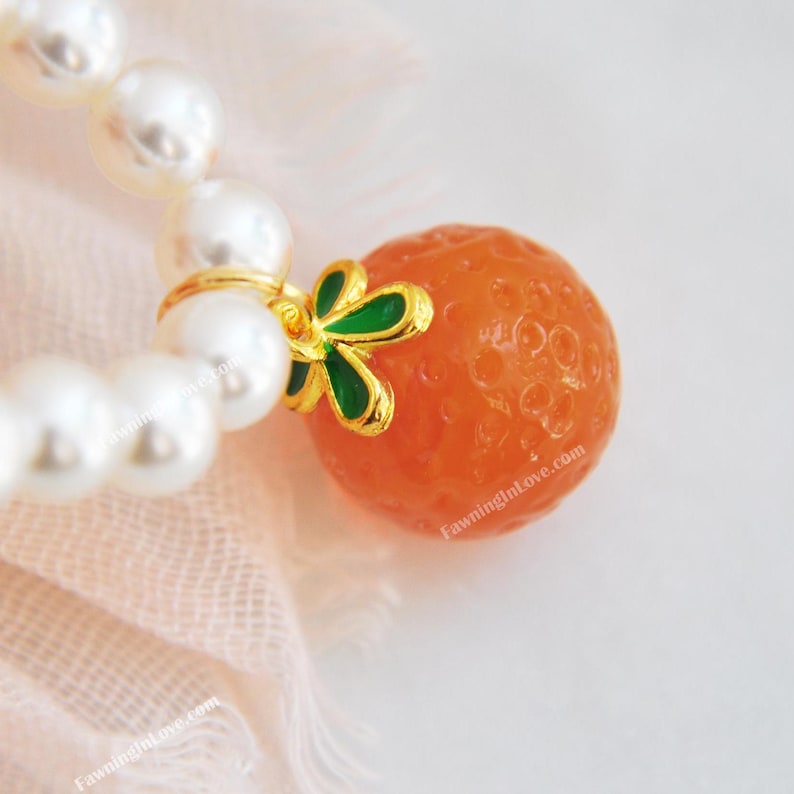 May include: A close-up of a pearl bracelet with a gold charm. The charm is an orange sphere with a gold and green accent. The bracelet is on a pink fabric.