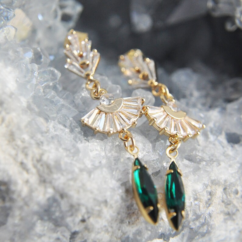 May include: A pair of gold-toned earrings with a fan-shaped design, adorned with clear crystals and emerald green teardrop-shaped gems. The earrings are displayed against a light blue, crystalline background.