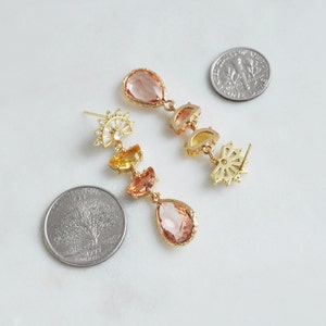 May include: Gold-toned drop earrings with peach, yellow, and orange-colored gemstones. The earrings feature a sunburst design and teardrop-shaped stones. The earrings are displayed on a white surface next to two coins.