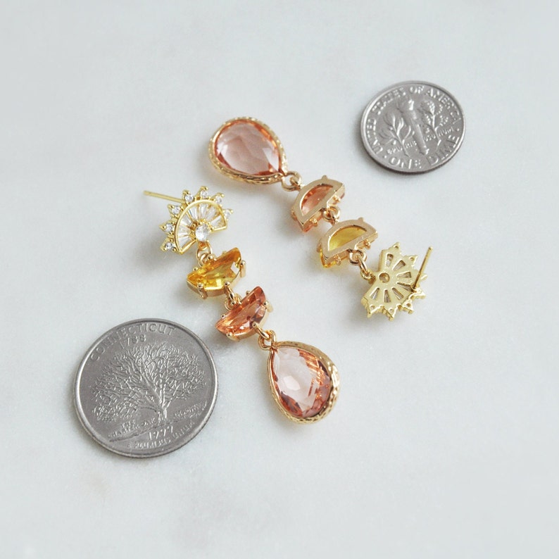 May include: Gold-toned drop earrings with peach, yellow, and orange-colored gemstones. The earrings feature a sunburst design and teardrop-shaped stones. The earrings are displayed on a white surface next to two coins.