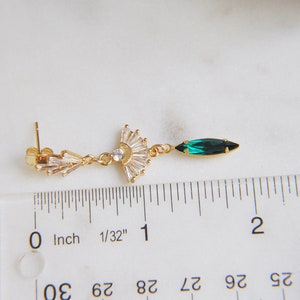 May include: A gold-toned earring with a fan-shaped design of clear crystals and a single emerald-green marquise-cut crystal. The earring is measured with a ruler in inches.