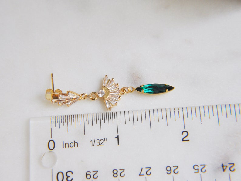 May include: A gold-toned earring with a fan-shaped design of clear crystals and a single emerald-green marquise-cut crystal. The earring is measured with a ruler in inches.