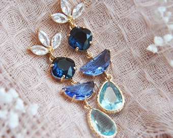 May include: Gold-toned dangle earrings featuring a cluster of clear, blue, and teal-colored gemstones. The earrings have a floral design at the top with clear stones, and the dangling portion includes various shapes of blue and teal gems.
