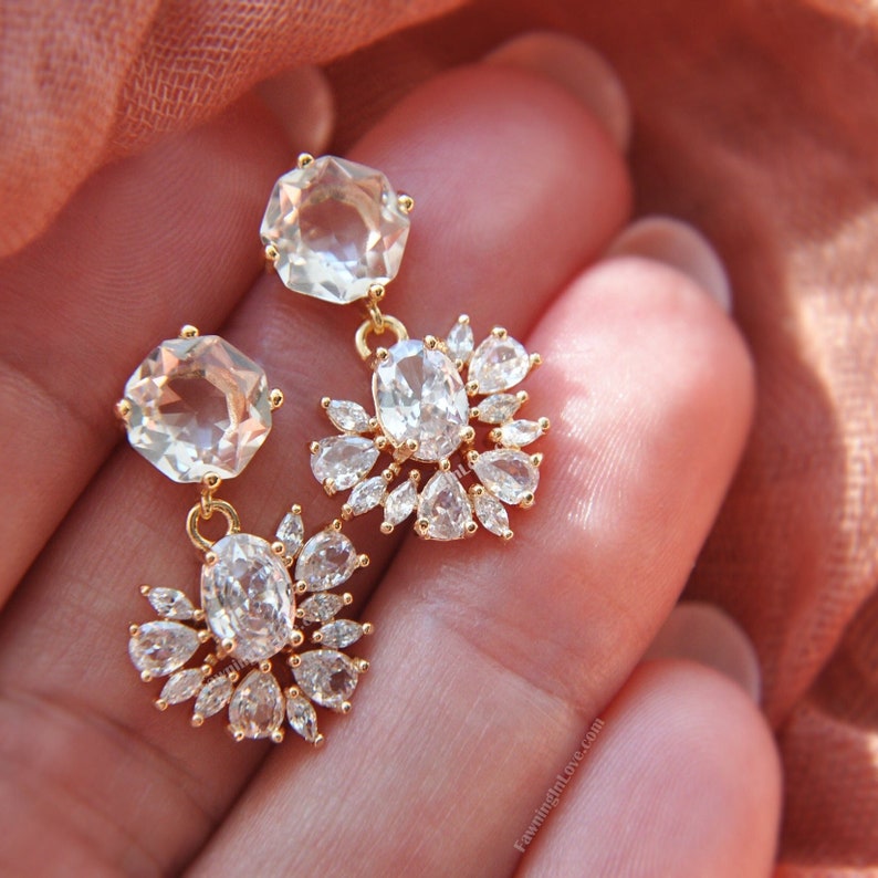 May include: Gold-toned drop earrings featuring large, faceted, light-colored gemstones. Each earring has a large, octagonal stone at the top, connected to a cluster of smaller, oval and teardrop-shaped stones. The earrings are designed for pierced ears.
