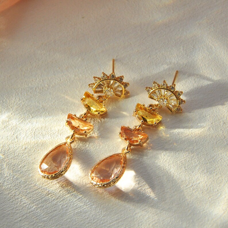 May include: A pair of gold-toned earrings with a sunburst design at the top. Each earring features yellow, orange, and peach-colored gemstone accents, culminating in a teardrop-shaped peach-colored stone.