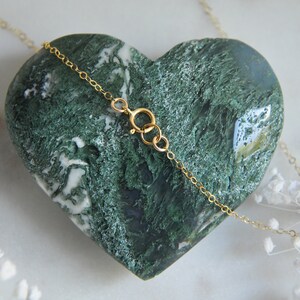 Puede incluir: Un delicado collar de cadena dorada con cierre de mosquet&oacute;n descansa sobre una piedra de &aacute;gata musgosa en forma de coraz&oacute;n. La piedra es de color verde oscuro con patrones blancos y verde claro. Flores blancas dispersas.