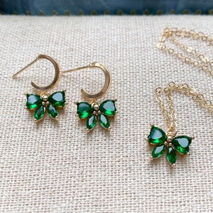 Puede incluir: Un conjunto de joyas de color dorado que incluye un par de pendientes de aro y un collar, cada uno adornado con un colgante en forma de lazo. Los lazos est&aacute;n adornados con piedras preciosas verdes vibrantes, creando un contraste llamativo con el oro.