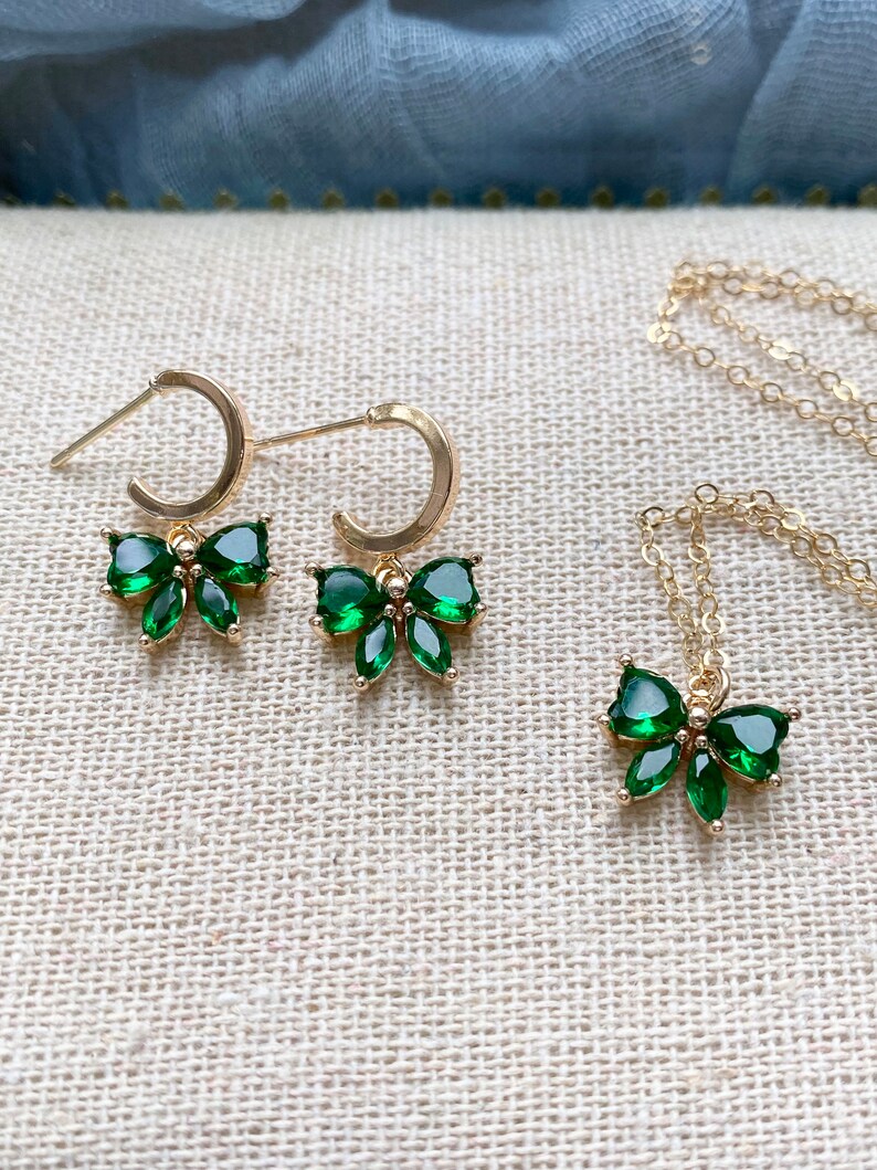 Puede incluir: Un conjunto de joyas de color dorado que incluye un par de pendientes de aro y un collar, cada uno adornado con un colgante en forma de lazo. Los lazos est&aacute;n adornados con piedras preciosas verdes vibrantes, creando un contraste llamativo con el oro.