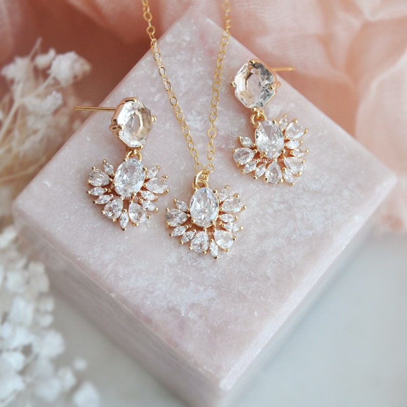 May include: A gold-toned jewelry set featuring a necklace and earrings. The necklace has a pendant with a central oval-shaped clear stone surrounded by smaller clear stones. The earrings have a similar design, with a large clear stone at the top and a cluster of smaller stones below.