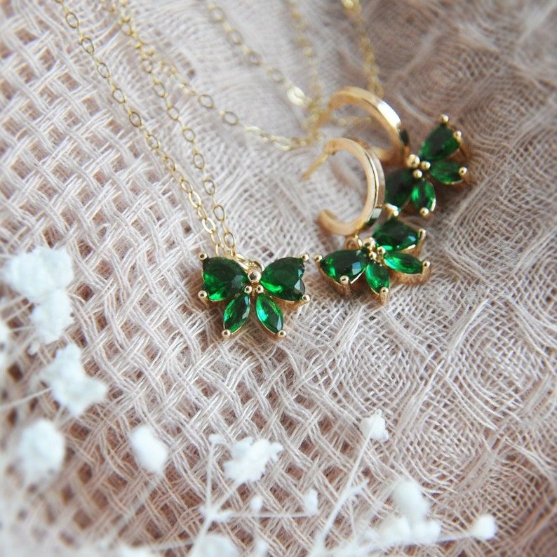 Puede incluir: Juego de joyas doradas que incluye un collar y pendientes. El collar tiene un colgante con detalles de piedras preciosas verdes en forma de mariposa. Los pendientes son de estilo aro con detalles de mariposa de piedras preciosas verdes a juego.