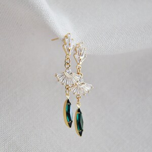 May include: Gold-toned drop earrings featuring a fan-shaped design with clear crystals and emerald green teardrop-shaped gems. The earrings have a post-back closure and are displayed against a white background.