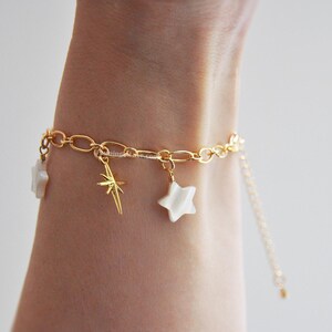May include: Gold-toned chain bracelet with star charms. The bracelet features a delicate chain design with three star-shaped charms, two white and one gold, and a gold starburst charm. The bracelet is shown on a person's wrist.