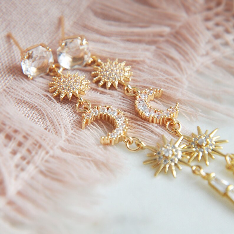 May include: Gold-toned celestial earrings featuring clear crystal studs, sunbursts, and crescent moons, all adorned with sparkling crystals. These dangle earrings are a stylish accessory.